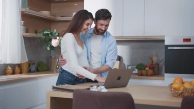 Kafkasyalı hamile, genç ve çekici bir kadın ve mutlu bir koca dizüstü bilgisayarda görüntülü sohbet ediyor. Haberleri ve ultrason fotoğrafını paylaşıyorlar. Gelecekteki ebeveynler. Mutfağa. Ev.