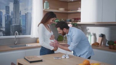 Beyaz bir aile. Hamile, genç, beyaz bir kadın kocasıyla gülerek hamilelik karnını gösteren bir çocuk bekliyor. Eğlenceli iletişim. Ebeveynler.