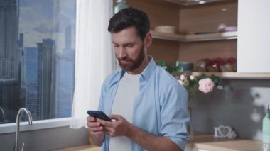 Bir adam akıllı telefonunu güzel şehir manzarası olan modern bir mutfakta dururken kullanıyor.