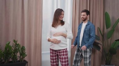 Hamile, genç ve neşeli bir kadın kocasıyla oturma odasında sarılıyor. Sevgi dolu bir aile. Mutluluk, neşe ve sağlık.