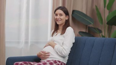 Beyaz hamile kadın kameraya neşeyle gülümsüyor, karnına nazikçe sarılıyor ve kanepeye oturuyor. Planlı hamilelik. Sağlık hizmetleri. Portreler.