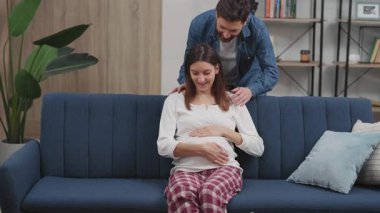 Kanepede oturan güzel karısını kucaklayan mutlu yakışıklı adam. Kocası oturma odasında olan sağlıklı, hamile bir kadın. Aile hayatı. Hamilelik.