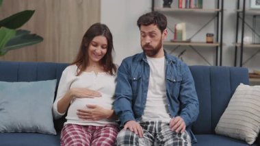 Hamilelik sorunları. Sinirli, korkmuş, sakallı genç bir adam kanepeye yaslanıyor ve mutlu hamile sevgilisinin yanına doğru kayıyor. Planlanmamış hamilelik.