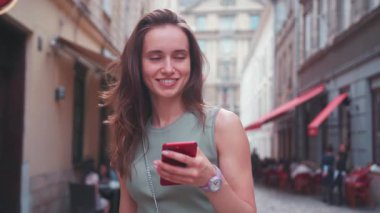Neşeli genç bir kadın, güzel bir şehir merkezinde akıllı telefonuyla mutlu mesut takılıyor.