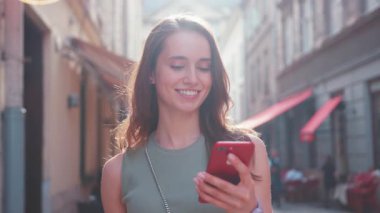 Neşeli genç bir kadın canlı bir şehir caddesini keşfederken parlak kırmızı akıllı telefonunu kullanmaktan mutluluk duyar.