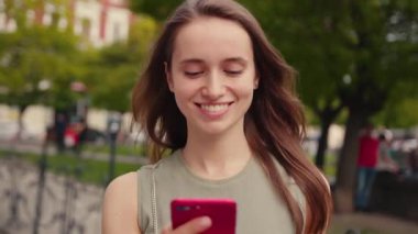 A joyful young woman happily smiling while using her smartphone outdoors, surrounded by vibrant greenery and nature