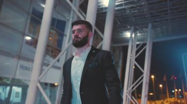 Şık takım elbiseli iyi giyimli bir adam gece canlı bir şehir bölgesinde, kendinden emin adımlarla yürüyor ve karizma yayıyor.