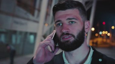 Şık bir genç adam, gece saatlerinde canlı şehir atmosferinin ortasında bir telefon görüşmesi yapıyor.
