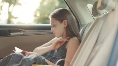 Genç ve güzel bir bayan arabanın arka koltuğunda oturmuş kitap okuyor, saçlarına dokunuyor. Güzel bir kızın görüntüsü. Şefkat. Gündüz, ulaşım, araç, ulaşım, otomobil, seyahat