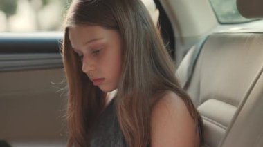 Güzel görünümlü küçük kız arka koltukta oturuyor, aşağı bakıyor. Kameraya bakıp gülümseyen tatlı bir gencin yakın plan çekimi. Otomobil. Günlük yaşam.
