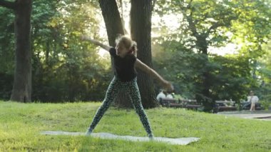 Beyaz tenli, spor elbiseli, dışarıda yoga yapan küçük bir kız. Esnek vücut. Formda ve sağlıklı. Yaz zamanı. Park et. Şehre. Şimdiki zaman.