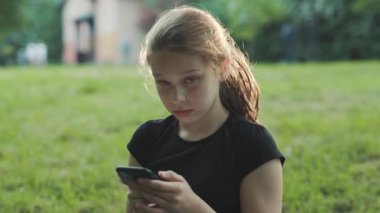 Siyah akıllı telefon kullanan at kuyruklu güzel beyaz bir kızın görüntüsü. Kameraya bakan güzel bir genç kızın portresi. Gülümse. Yeşil arka plan. Açık havada..