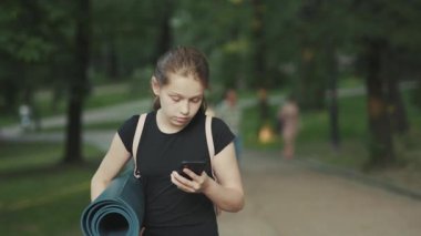 NEW YORK - 19 Nisan 2019: yoga minderli hoş beyaz kız parkta yürüyor, akıllı telefon ekranına bakıyor, mesajlaşıyor. Yaz zamanı. Şehre. Günlük hayat. Açık hava..