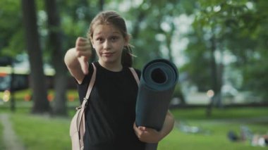 Üzücü hayal kırıklığı, spor kıyafetleri ve yoga minderi giymiş harika genç bir kadının kameraya bakması ve baş parmağını kaldırması. Açık havada. Yaz zamanı. Yeşil Park.