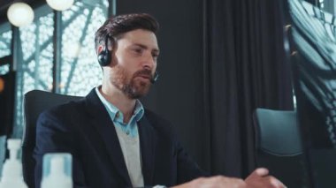 Yakın çekim genç adam iş için kulaklık kullanıyor bilgisayarla konuşuyor modern ofis programcısı gülümsüyor monitörde çalışan başarılı iş adamına bakıyor yavaş çekim