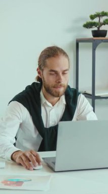 Bu videoda, sakallı bir adam bir masada oturuyor ve dizüstü bilgisayarı üzerinde çalışıyor. Heyecan içinde ellerini kaldırır, beyaz gömlek ve yeşil kazak giyer, başarının neşeli anını kutlar.