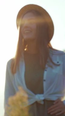 Hasır şapkalı ve beyaz gömlekli neşeli bir kadın güneş ışığında güneşleniyor, gülümsüyor ve pozitif ışıldıyor. Neşeli bir şekilde yüzünü kapatıyor, yazları kaygısız bir ruh sergiliyor.