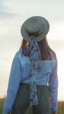 Şık hasır şapkalı bir kadın güneşli bir tarlada uzun otların arasında narin bir eşarp takıyor. Bu video doğanın güzelliğini yakalar.