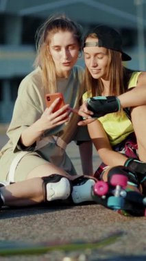 İki genç kadın yerde rahatça oturuyorlar, cep telefonlarını yakından izliyorlar, birbirleriyle iletişim kurarken eğlenceli bir anı paylaşıyorlar.