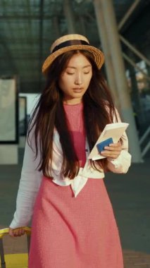 Renkli pembe elbiseli ve şık şapkalı genç bir kadın bavuluyla güvenle duruyor, yolculuğa hazır.