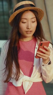 Pembe elbisesi ve hasır şapkası olan genç bir kadın telefonunu kontrol ediyor ve son moda bir yaz havası yayıyor.