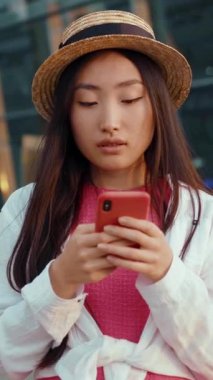 Şık bir hasır şapka giyen genç bir kadın canlı bir şehir manzarasının ortasında akıllı telefonuyla derinden meşgul.