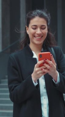 Modayı takip eden genç bir kadın açık havada hareketli bir şehir ortamında akıllı telefonuyla aktif olarak meşgul oluyor.