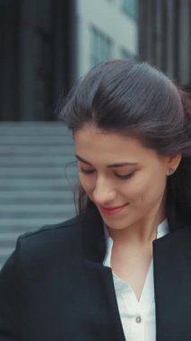 Kendine güvenen genç bir kadın, modern bir şehir ortamında gülümseyerek ve çevresinin tadını çıkarırken dışarıda duruyor.