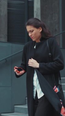 Şık siyah bir ceket giymiş bir kadın akıllı telefonunu tutarken düşünceli bir şekilde modern yaşam tarzını yansıtıyor.