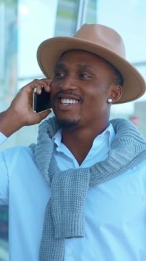 Şık bir genç adam şık bir şapka ve şık bir süveter giyerek akıllı telefonuyla konuşur.