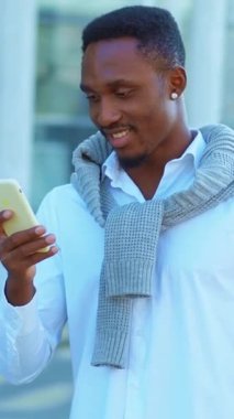 Aydınlık ve güneşli bir günde şık giyinmiş neşeli bir adam açık havada telefonuna mutlu bir şekilde bakar.