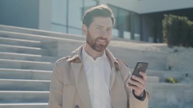 Güneş ışığında çekici yüzlü şık, ceketli, sırıtan bir adam akıllı telefon standı kullanıyor. Kapatın. Yavaş çekim