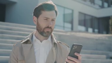 Ciddi çekici yüz stili palto giyen sakallı adam güneş ışığında gölün kenarında akıllı telefon standı kullanıyor. Kapatın. Yavaş çekim