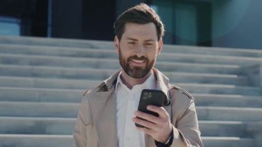 Portre genç çekici yüz stil adam ceketli sakallı arka plandaki merdivenlerde akıllı telefon gülümseme standı kullanıyor. Kapatın. Yavaş çekim