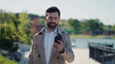 Palto giyen yakışıklı bir işadamı göl kenarındaki parkta akıllı telefon kullanıyor. Gülümseyerek etrafına bak. Kapatın. Yavaş çekim