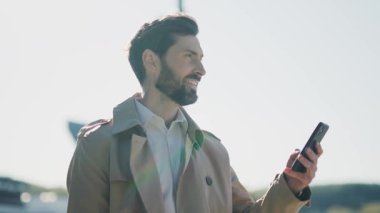 Portre stili, genç sakallı, ceketli yakışıklı adam sokakta akıllı telefon standı kullanıyor. Gülümseyerek mutlu ol. Kapatın. Yavaş çekim