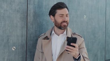 Portre stili ceketli yakışıklı adam caddedeki akıllı telefon standında daktilo kullanıyor. Gülümseyerek mutlu ol. Kapatın. Yavaş çekim