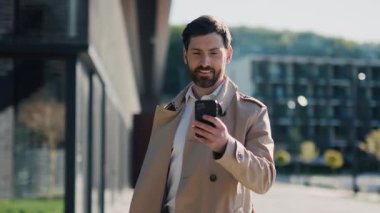 Park yakınlarında şık, ceketli yakışıklı bir adam güneş ışığında akıllı telefon kullanıyor. Gülümseyerek mutlu ol. Yavaş çekim