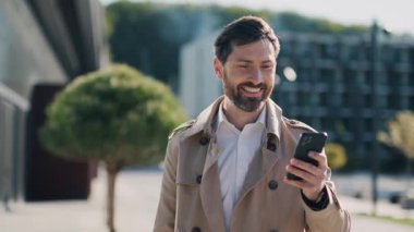 Paltolu yakışıklı adam sokakta akıllı telefon kullanıyor. Gülümseyerek mutlu ol. Yürüyorum. Yavaş çekim
