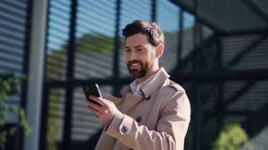 Paltolu şık genç çekici adam sokakta akıllı telefon standı kullanıyor. Gülümseyerek mutlu ol. Kapatın. Yavaş çekim
