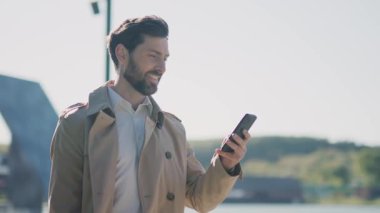 Çekici, yakışıklı, şık montlu adam sokakta akıllı telefon kullanıyor. Gülümseyerek mutlu ol. Yürüyorum. Kapatın. Yavaş çekim