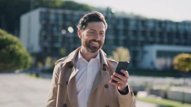 Paltoyla gülümseyen şık, yakışıklı adam sokakta akıllı telefon kullanıyor. Mutlu ol. Yürüyorum. Kapatın. Yavaş çekim