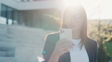 Şehir merkezinde güneş ışığında telefonu kullanan çekici genç bir kadın göl kenarındaki parka bakıyor. Cep telefonu olan bir portre kızı. Yavaş çekim