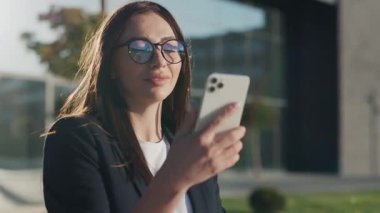 Gün ışığında, şehirde gülümseyen modern bir akıllı telefon standı kullanarak gözlüklü genç ve güzel bir iş kadını. Yavaş çekim