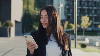 Şehir merkezinde gün ışığında telefonla yürüyen çekici bir kadın portresi. Etrafına bak ve mutlu ol. Yavaş çekim