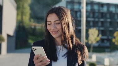 Güler yüzlü genç ve çekici bir kadın şehir merkezinde gün ışığında telefonla yürürken etrafına bak ve mutlu ol. Cep telefonu olan bir portre kızı. Yavaş çekim