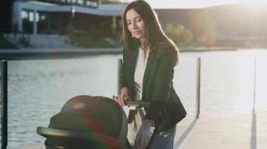 Çekici genç bir kadın göl kenarındaki güneşli parkta bebek arabasıyla dikiliyor. Etrafa bakıp gülümsüyorum. Annelik. Kapatın. Yavaş çekim