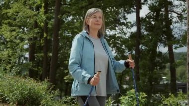 Çekici, gülümseyen, yaşlı, beyaz bir kadının İskandinav bastonlarıyla yaptığı yürüyüş sırasında doğada vakit geçirmesinin ön görüntüsü. Ağır çekim. Tatil köyü kavramı. Tatil, lesuire, açık hava. Tek başıma. Sağlıklı bir yaşam tarzı. Portre