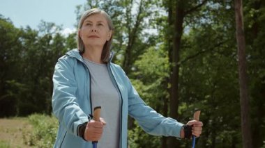 Sopalarıyla yürüyen muhteşem, sportif yaşlı bir kadının görüntüsü. Spor elbiseli çekici bir kadının portresi doğada İskandinav yürüyüşü yapıyor. Otel kompleksi. Sağlıklı eğitim, yaşam tarzı. Rahatlatıcı, huzurlu, dinlendirici, açık havada