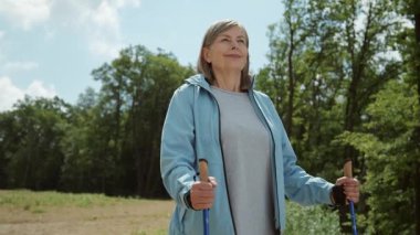 İskandinav yürüyüş çubuklarıyla güzel, kıdemli, beyaz bir kadın. Uzağa bakan spor elbiseli yaşlı kadın portresi. Tarla, arka planda ağaçlar. Yaz zamanı.